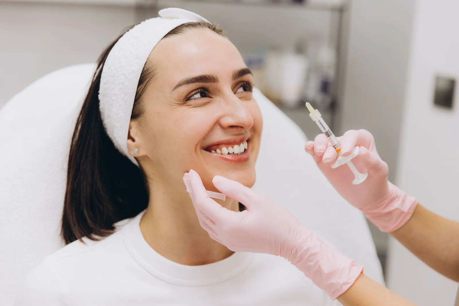 Smiling patient in a treatment chair as a provider in pink gloves holds a small syringe beside her face