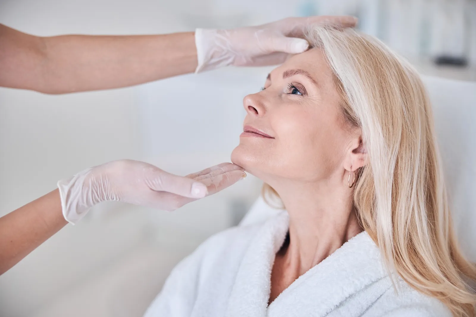 Smiling woman in a white robe as a provider's gloved hands gently position her face for an assessment