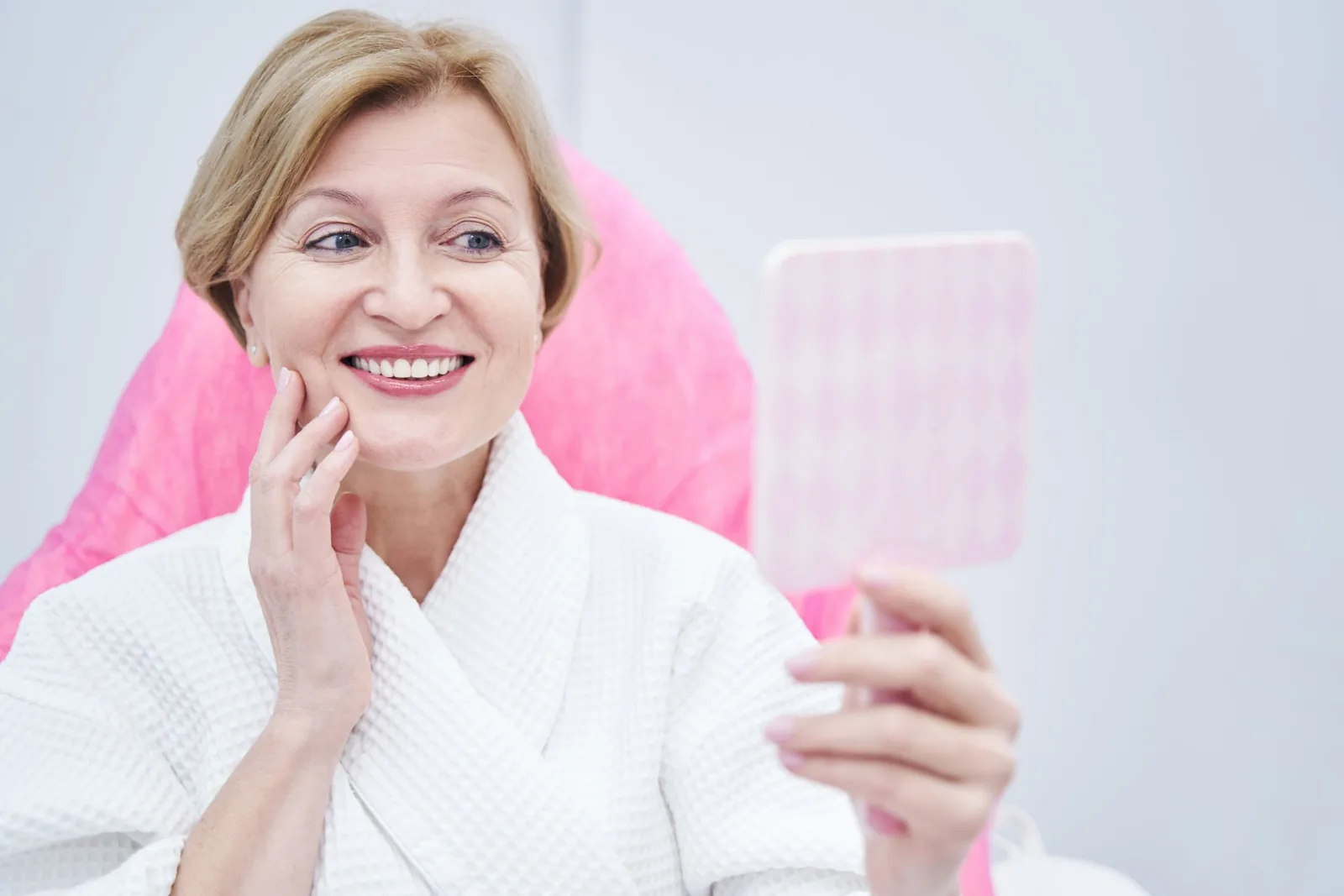 Woman in a treatment robe smiling as she checks her cheeks in a hand mirror during a facial volume consultation in Orlando