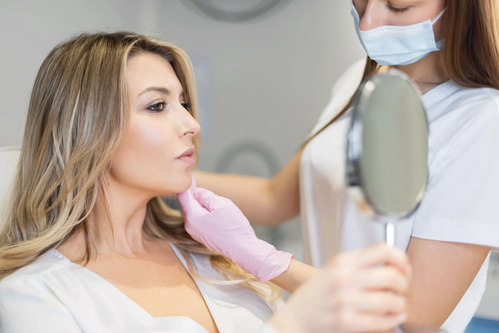 Provider supporting a patient's chin with a gloved hand while she studies her jawline in a hand mirror