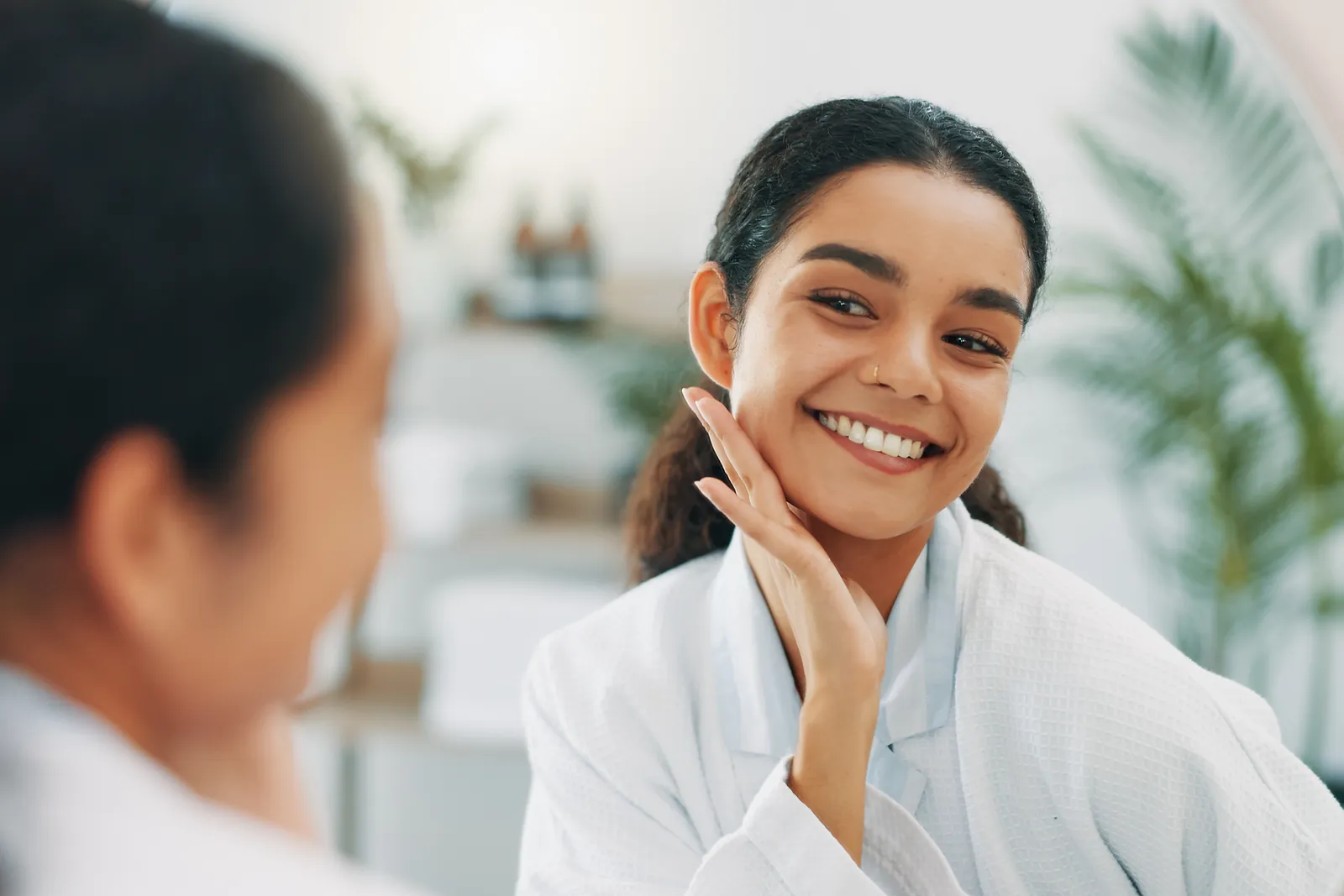 Smiling woman in a bathrobe touching her jaw as she looks at her reflection in a round mirror