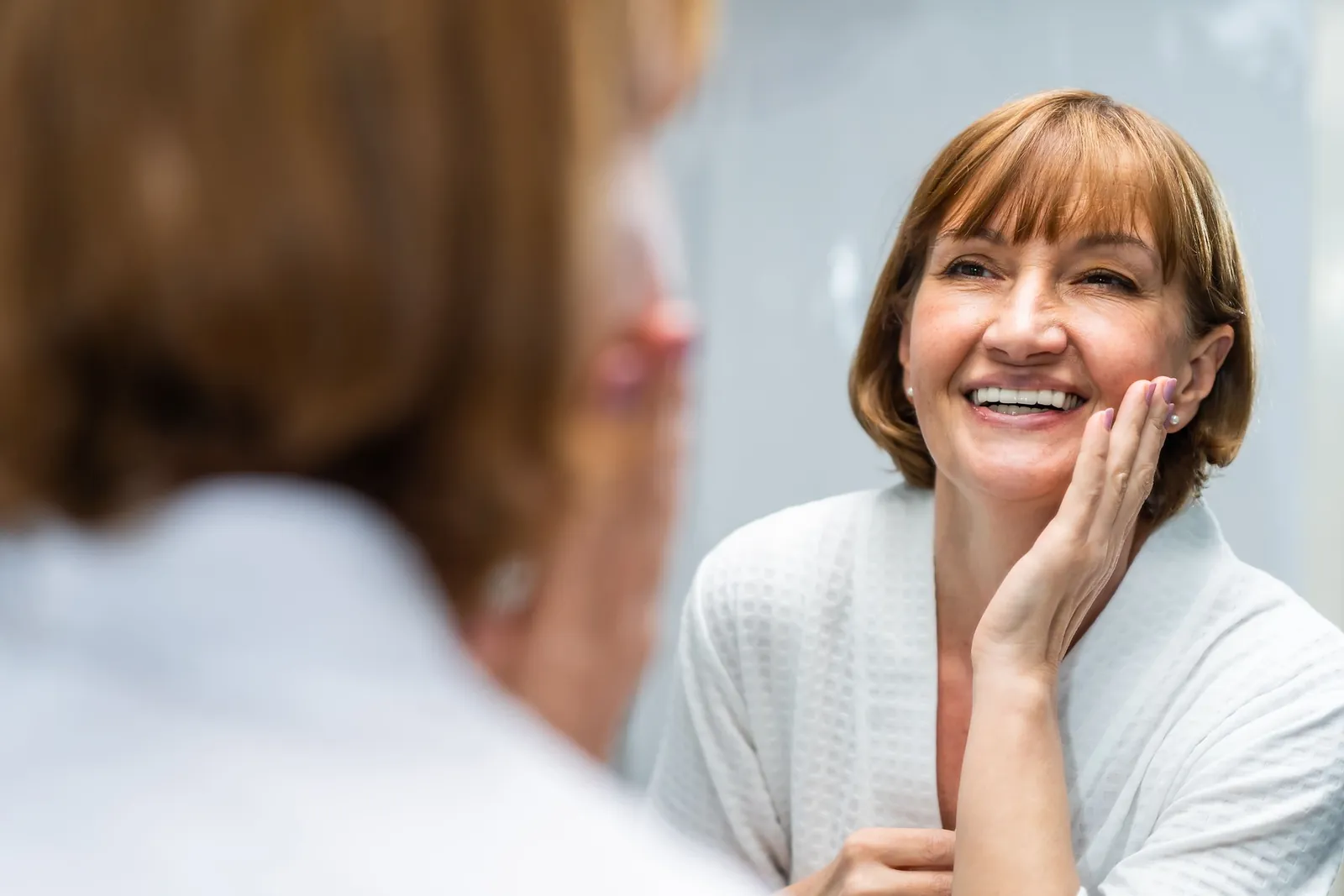 Smiling mature woman in a bathrobe resting her hand on her cheek as she looks in the mirror
